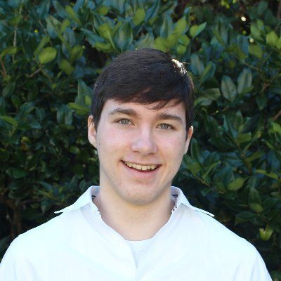 A headshot of a young man with short dark brown hair and a white shirt, smiling in front of a backdrop of green leaves