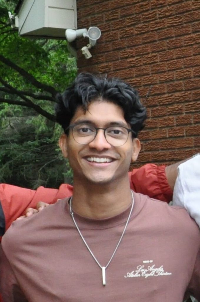 A headshot of a man with short black hair, glasses, and a brown T-shirt in front of a brick wall.